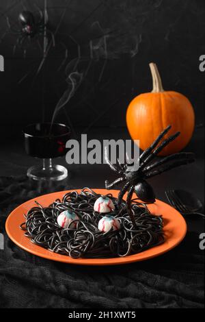 Halloween lustige italienische schwarze Pasta Tintenfisch Tinte verzierte Spinnen in orange Platte mit Rauchtrank auf schwarzem Hintergrund. Horror-Essen für Party. Vertikal Stockfoto