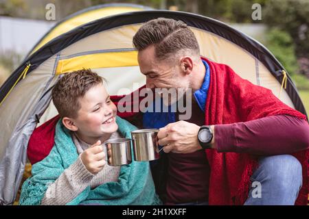 Kaukasischer Vater und Sohn lächeln, während sie ihre Kaffeetassen in einem Zelt im Garten toasten Stockfoto