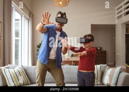 Kaukasischer Vater und Sohn mit vr-Headsets gestikulieren, während sie zu Hause im Wohnzimmer stehen Stockfoto