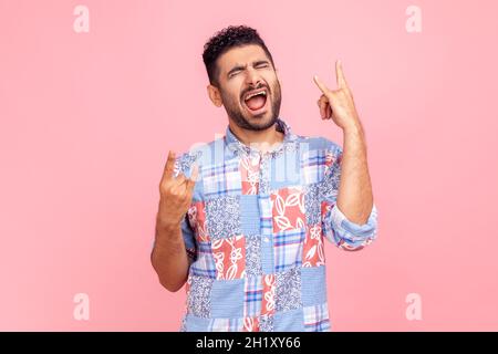 Rock 'n' Roll. Lustiger junger erwachsener bärtiger Mann mit blauem Hemd im lässigen Stil, der mit Rock-Singen steht, schreiend und aufgeregt im Gesichtsausdruck schreit. Innenaufnahme des Studios isoliert auf rosa Hintergrund. Stockfoto