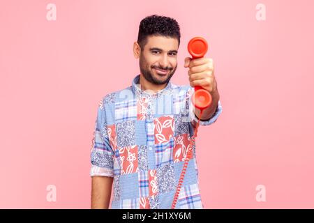 Brunet-Mann mit Bart trägt ein blaues Hemd im lässigen Stil, hält das Festnetztelefon und zeigt es mit einem Lächeln auf die Kamera. Innenaufnahme des Studios isoliert auf rosa Hintergrund. Stockfoto