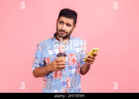 Porträt eines gutaussehenden nachdenklichen jungen erwachsenen Blogger Mann Bart in blauem Hemd stehend mit Kaffee und Chat am Telefon, wegschauen. Innenaufnahme des Studios isoliert auf rosa Hintergrund. Stockfoto