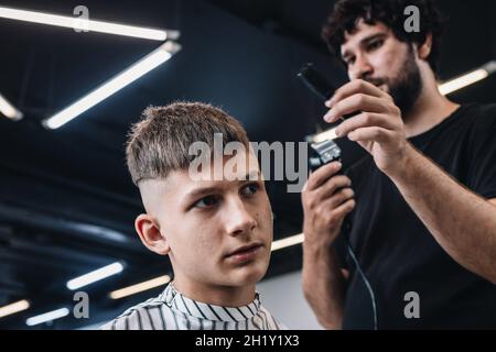 Stilvolle bärtige Brünette Friseur macht einen modischen Haarschnitt zu einem jungen Kerl. Menschen bei der Arbeit. Konzept für kleine Unternehmen oder Dienstleistungen. Professioneller Haarschnitt A Stockfoto