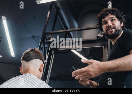 Stilvolle bärtige Brünette Friseur macht einen modischen Haarschnitt zu einem jungen Kerl. Menschen bei der Arbeit. Konzept für kleine Unternehmen oder Dienstleistungen. Professioneller Haarschnitt A Stockfoto