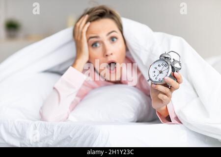 Overleeping. Porträt einer schockierten Frau, die die Uhr im Bett hält Stockfoto