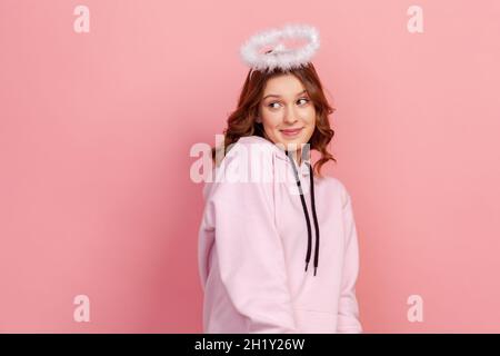 Porträt eines schüchternen lockigen Teenagers im Kapuzenpullover, der mit Halo auf dem Kopf steht und mit einem Lächeln und einem träumenden Gesicht wegschaut, Engel. Innenaufnahme des Studios isoliert auf rosa Hintergrund Stockfoto