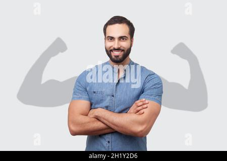 Erfolgreicher, positiver bärtiger Mann in blauem Hemd, der die Hände auf der Brust zusammenfaltete und mit einem toothy Lächeln auf die Kamera schaute und starke, große Arme im Schatten zeigte. Innenaufnahme des Studios isoliert auf grauem Hintergrund Stockfoto