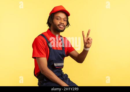 Porträt eines attraktiven bärtigen Handwerkers mit Dreadlocks in blauer Uniform, roter Visierkappe und T-Shirt in Pose, Blick auf die Kamera, mit V-Zeichen. Innenaufnahme des Studios isoliert auf gelbem Hintergrund. Stockfoto