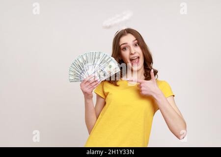 Porträt eines fröhlichen, dunkelhaarigen Teenagers im lässigen T-Shirt mit Halo am Kopf, der mit dem Finger auf eine Reihe von Dollar-Banknoten zeigt. Innenaufnahme des Studios isoliert auf grauem Hintergrund. Stockfoto