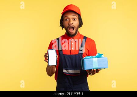 Porträt des überraschten Kuriers in blauer Uniform halten und zeigen Geschenkboxen und Smartphone mit leerem Display, Auftrag Anwendung. Innenaufnahme des Studios isoliert auf gelbem Hintergrund. Stockfoto