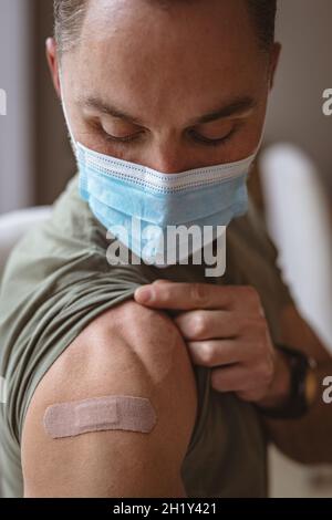 Kaukasischer Mann mit Gesichtsmaske, der zu Hause seine geimpfte Schulter zeigt Stockfoto