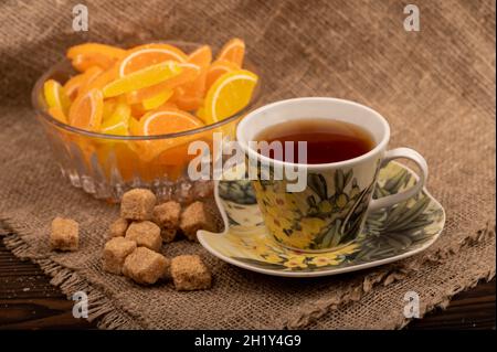 Bunte, geformte Marmeladenscheiben in einer Glasvase, Stücke braunen Rohrzuckers und eine Tasse Tee auf einer Untertasse, Nahaufnahme, selektiver Fokus Stockfoto