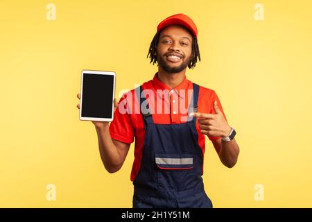 Porträt eines zufriedenen bärtigen Handyman trägt Overalls hält Tablet mit leerem Bildschirm für Werbung Industrie Service, zeigt auf Display. Innenaufnahme des Studios isoliert auf gelbem Hintergrund. Stockfoto