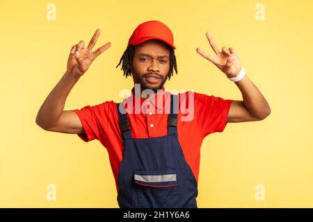 Positiver Handwerker mit Bart in Overalls Blick auf die Kamera, zeigt V-Zeichen, arbeitet in der Service-Industrie, Kurierdienst, Housekeeping Wartung. Innenaufnahme des Studios isoliert auf gelbem Hintergrund. Stockfoto