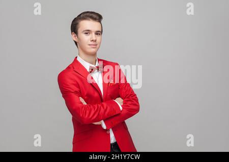 Portrait eines trendigen, gutaussehenden Gentleman mit stilvoller Frisur in rotem Smoking und Fliege, die mit gekreuzten Händen und positivem, ruhigem Ausdruck steht Stockfoto