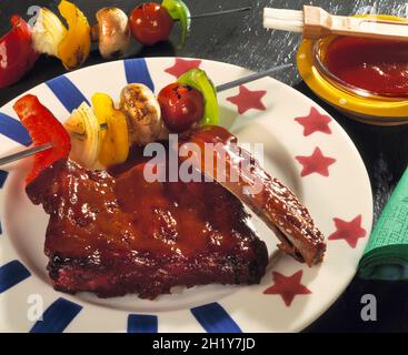 BBQ Spare Ribs mit Sauce & Spieß mit gebratenem Gemüse rote Paprika Tomate auf Sterne & Streifen Teller auf glänzendem Schiefer mit roter Sauce & Basting Pinsel Stockfoto