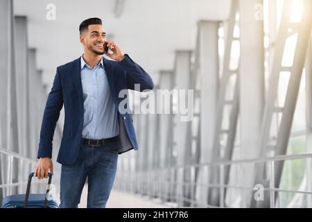 Junger Geschäftsmann Aus Dem Nahen Osten, Der Mit Dem Koffer Im Flughafen Unterwegs Ist Und Sich Mit Dem Handy Unterhält Stockfoto