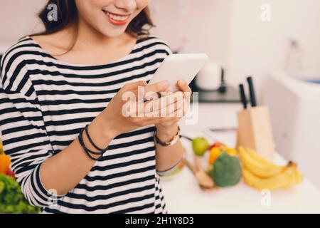 Attraktives junges Mädchen, das mit dem Mobiltelefon beschäftigt ist oder Updates auf dem Smartphone sucht, während es in der Küche eine Schürze trägt Stockfoto