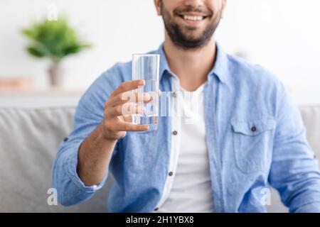 Lächelnder Kaukasier mittleren Alters mit Bart sitzt auf dem Sofa und hält Glas mit sauberem stillem Wasser Stockfoto