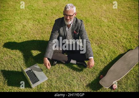Ein grauhaariger Geschäftsmann sitzt auf dem Gras und meditiert Stockfoto
