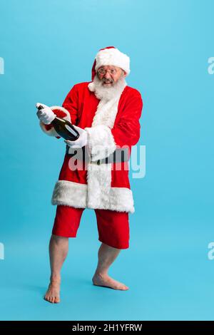 Ganzkörperportrait eines älteren Mannes, der das Weihnachtsmann-Kostüm trägt und eine Champagnerflasche auf blauem Hintergrund öffnet. Feiertagsfeier Stockfoto