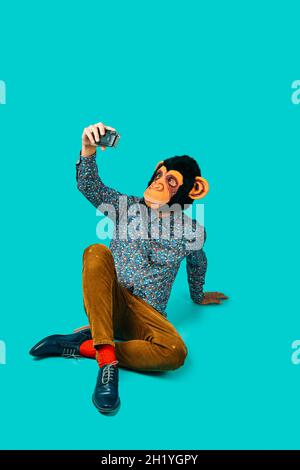 Ein junger Mann, der eine Affenmaske trägt, macht ein Selfie mit einer Retro-Filmkamera, sitzt auf einem blauen Hintergrund mit etwas leerem Platz oben Stockfoto