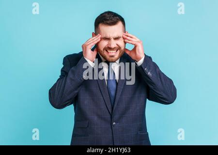 Unglücklich frustrierter bärtiger Mann mit offiziellem Anzug massiert Schläfen Gefühl Kopfschmerzen, leiden Migräne oder Bluthochdruck. Innenaufnahme des Studios isoliert auf blauem Hintergrund. Stockfoto