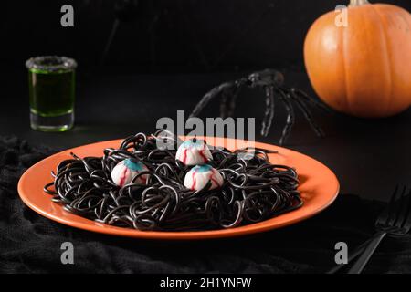 Halloween italienische schwarze Pasta Tintenfisch Tinte verzierte Spinnen, Augen in orange Teller mit Trank auf schwarzem Tisch. Horror lustiges Essen für Party. Nahaufnahme. Stockfoto