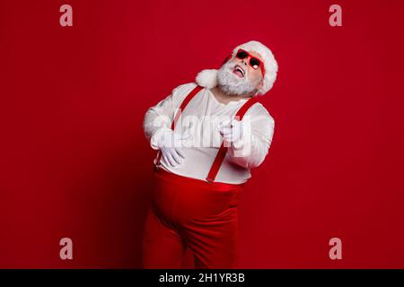 Foto von verrückten lustigen übergewichtigen Mann lachen direkten Finger tragen Sie santa Hut Kostüm isoliert roten Hintergrund Stockfoto