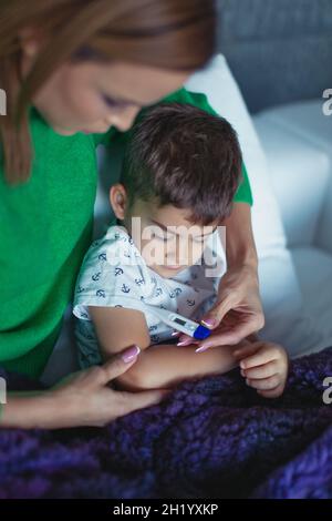 Junge kaukasische Mutter nimmt Temperatur unter Arm für kleinen Sohn zu Hause Stockfoto