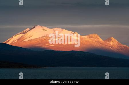 Episches Sonnenaufgangslicht beleuchtet felsigen, gletscherbedeckten Gipfel mit dunklem bewölktem Himmel im Hintergrund. Ahkka Peak im wunderschönen goldenen Stundenlicht, Stora Stockfoto