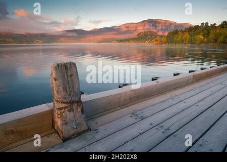 Die ersten Sonnenstrahlen treffen auf den Berg über dem See mit Holzsteg im Vordergrund. Rotes Licht auf einem Berggipfel. Lulep Gierkav Berg über Lulealven See in Stockfoto