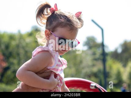 Kleines schönes Mädchen in Sonnenschutzbrille mit Zöpfen auf dem Spielplatz Stockfoto