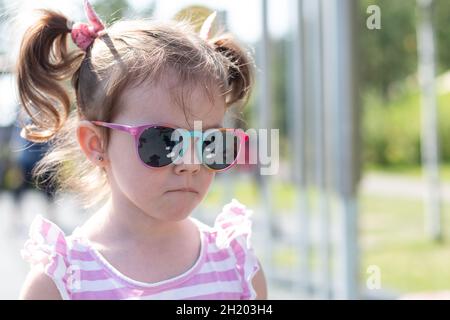 Kleine schöne traurige Mädchen in Sonnenschutzbrille mit Zöpfen steht auf der Straße Stockfoto