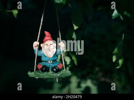 Garten-Zwerg, der auf einer Schaukel sitzt, die an einem Baum hängt, draußen im Hinterhof. Gartenarbeit Hintergrund oder Gartendekoration Hintergrund. Schweres Licht und Sha Stockfoto