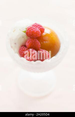 Dessertschüssel aus Milchglas mit einer Kugel Vanilleeis, einem pochierten ganzen Pfirsich und frisch gepflückten Himbeeren Stockfoto