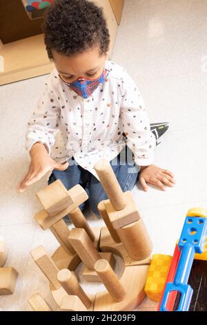 Bildung Vorschule 3-4-jähriger Junge, der im Blockbereich spielt und Holzklotz auf den Turm legt, den er baute, und Gesichtsmaske trägt, um vor Covid-19 zu schützen Stockfoto