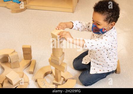 Bildung Vorschule 3-4-jähriger Junge, der im Blockbereich spielt und Holzklotz auf den Turm legt, den er baute, und Gesichtsmaske trägt, um vor Covid-19 zu schützen Stockfoto