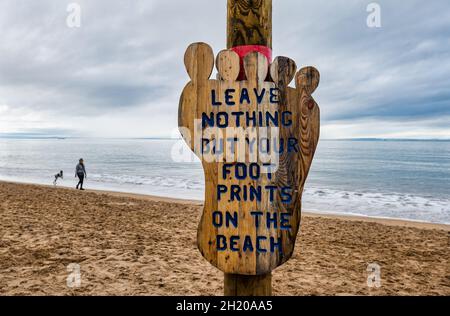 Gullane Bents, East Lothian, Schottland, Großbritannien, 19. Oktober 2021. Wetter in Großbritannien: Trüber, wolkiger Tag, an dem Himmel und Meer ineinander verschmelzen, während eine Frau und ein Hund an einem ansonsten menschenleeren Strand entlang des Sandufers spazieren gehen, mit einem hölzernen Umweltschild, das den Besuchern rät, nichts als Fußabdrücke am Strand zu hinterlassen Stockfoto