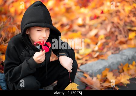 Der Junge, der eine schwarze Kapuzenjacke trägt, hält ein Herbstblatt in der Hand. Stockfoto