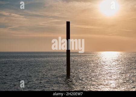 Große, rostige Stahlbausäule im blauen Meer mit ruhigen Wellen bei Sonnenuntergang, Sonnenlicht, das sich auf die Meerwasseroberfläche reflektiert. Vertikal verlassene Gebäudeteil Stockfoto