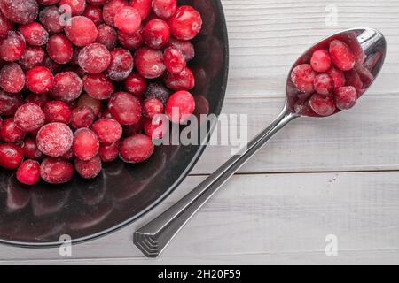 Frisch gefrorene rote Bio-Preiselbeeren mit einem Metalllöffel und einem schwarzen Teller, auf einem Holztisch, Nahaufnahme, Draufsicht. Stockfoto