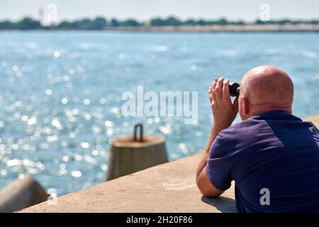 Älterer kahler Mann in T-Short, der am Pier steht und im Sommer durch ein Fernglas auf die blaue Ostsee blickt. Hände halten und fokussieren. Selektiver Fokus, Stockfoto