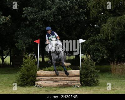 Frau, die auf einem grauen Pferd reitet und über einen Zaun mit dunklem Hintergrund in England springt Stockfoto