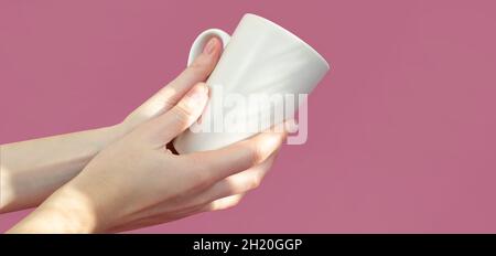 Weibliche Hände halten eine weiße Tasse auf einem rosa Hintergrund. Konzept für Food Drink. Speicherplatz kopieren. Stockfoto