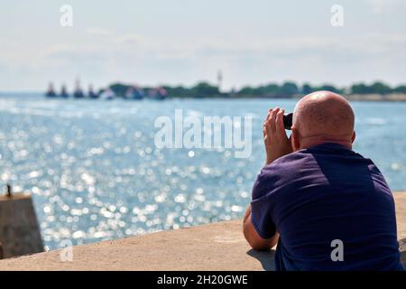 Älterer kahler Mann in T-Short, der während der Marineparade an der blauen Ostsee im Sommer auf dem Pier steht und durch ein Fernglas auf Kriegsschiffe blickt. Hände Holdi Stockfoto