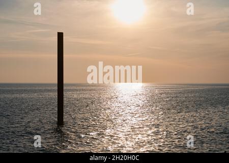 Große, rostige Stahlbausäule im blauen Meer mit ruhigen Wellen bei Sonnenuntergang, Sonnenlicht, das sich auf die Meerwasseroberfläche reflektiert. Vertikal verlassene Gebäudeteil Stockfoto