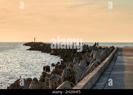 Lange Fußgängerpier mit Menschen, die in Europa bei Sonnenuntergang spazieren gehen. Sommerurlaub, Strandatmosphäre. Betonbrecher und blaues ruhiges Meer, b Stockfoto