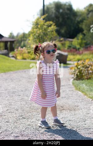 In der Mitte des Parks steht ein müdes kleines Mädchen mit Sonnenbrillen und einem gestreiften Kleid mit Zöpfen Stockfoto