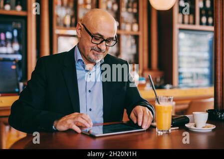 Bärtiger Senior in Anzug mit Tablet, während er in der Cafeteria saß. An der Rezeption gab es Saft und Kaffee. Stockfoto